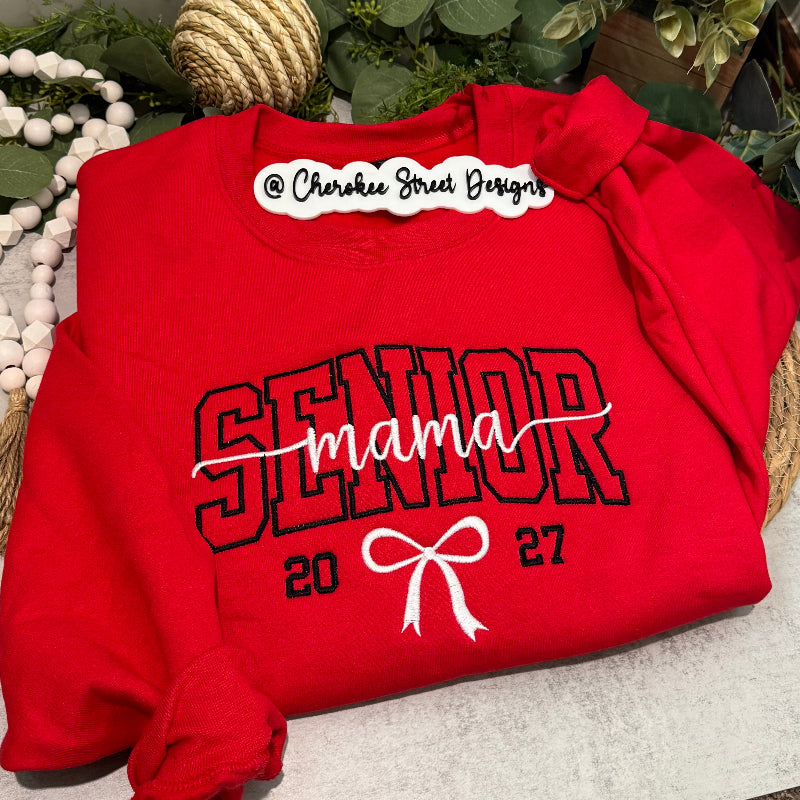 A folded red sweatshirt with 'SENIOR mama 2022' printed in black and white, styled on a neutral surface with decorative rope, greenery, and white beads.