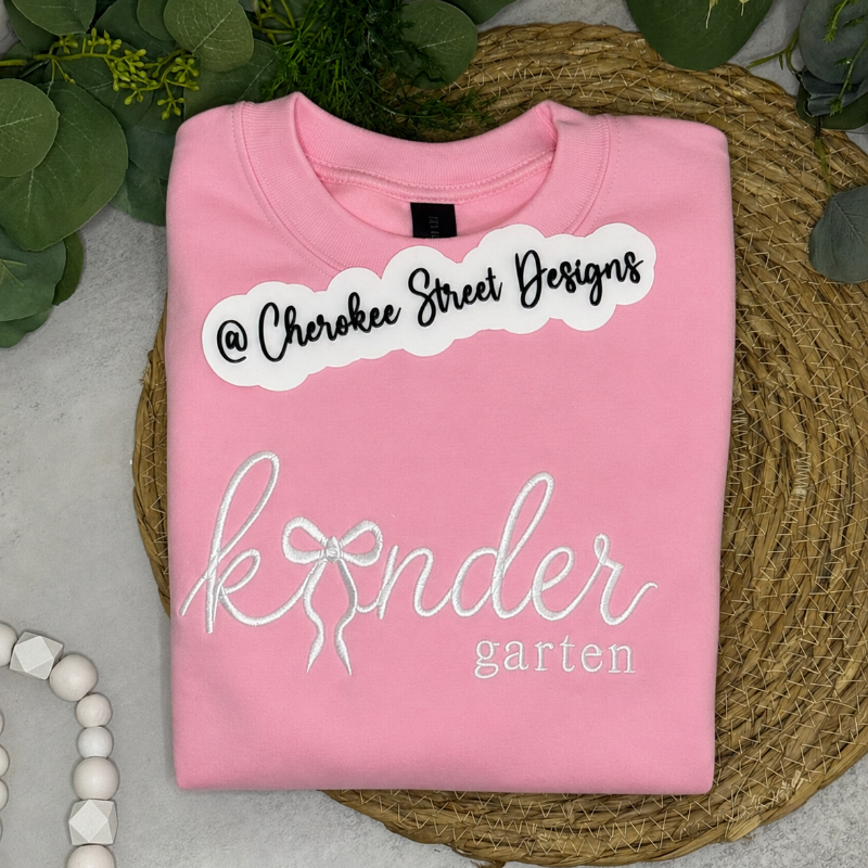 A folded pink crewneck sweatshirt with custom white and black graphic text reading  and 'kinder garden' with a bow illustration, 