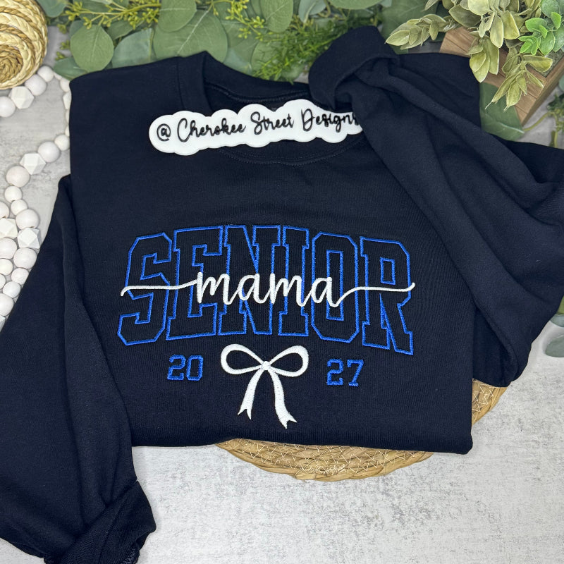 A navy blue sweatshirt laid flat on a light surface with decorative greenery and woven props around it. The sweatshirt has a large white and blue 'SENIOR mama 2027' graphic on the front, with a white script logo near the collar reading Cherokee Street Designs.
