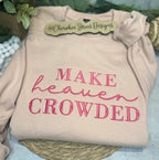 Beige sweatshirt with pink text 'MAKE heaven CROWDED' on a textured surface with greenery.