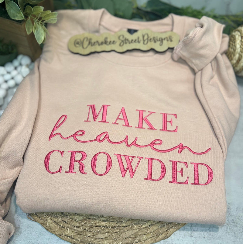 Beige sweatshirt with pink text 'MAKE heaven CROWDED' on a textured surface with greenery.