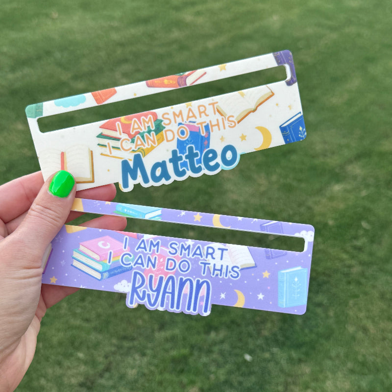 Two colorful plastic book dividers held in a hand against a green grass background. One is white with blue and orange graphics and the name 'Matteo', and the other is purple with blue and white graphics and the name 'Ryann'.