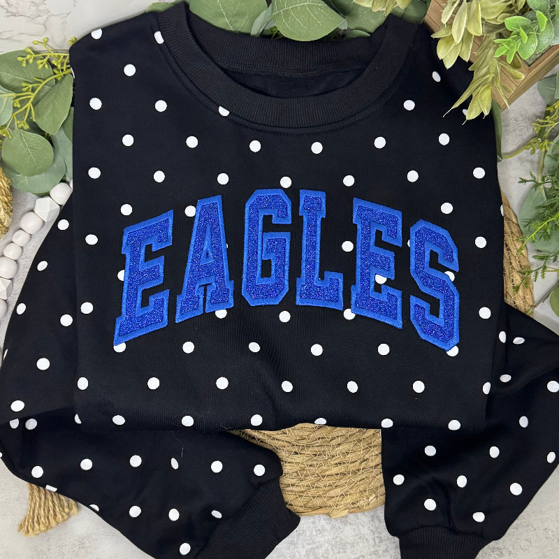 A black crewneck sweatshirt with white polka dots and 'EAGLES' embroidered in blue across the chest, styled with green leaves and a rope on a light stone surface.