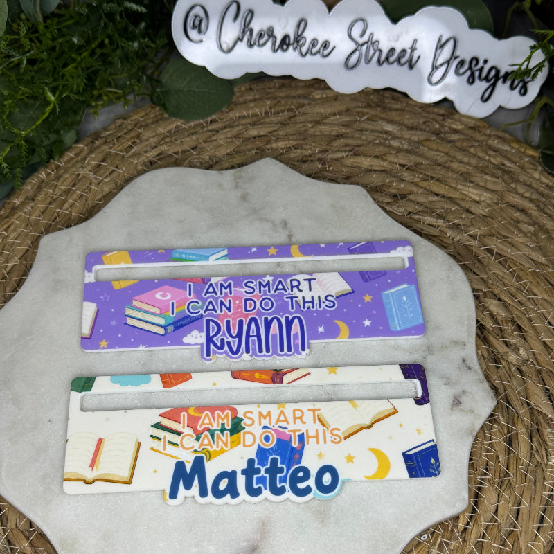 Two personalized book-themed nameplates displayed on a white scalloped stone plate, with a woven mat underneath and a sign reading '@ Cherokee Street Designs' in the background.