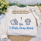 White sweatshirt with 'T-Ball Grandma' text and graphics on a textured surface with greenery.