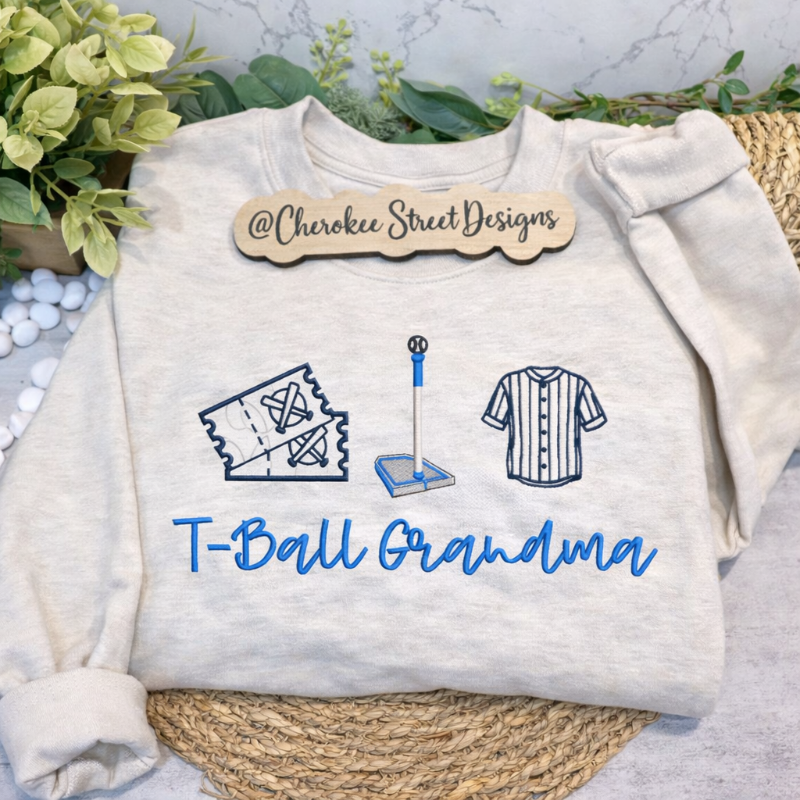 White sweatshirt with 'T-Ball Grandma' text and graphics on a textured surface with greenery.