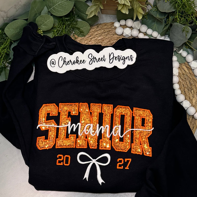 A black long-sleeve sweatshirt laid flat  The sweatshirt has a large orange glitter-style 'SENIOR' graphic, white script 'mama' across the center, white bow graphic, and orange '2027' text.