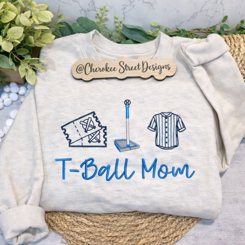 White sweatshirt with 'T-Ball Mom' text and graphics on a textured surface with greenery.