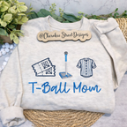 White sweatshirt with 'T-Ball Mom' text and graphics on a textured surface with greenery.