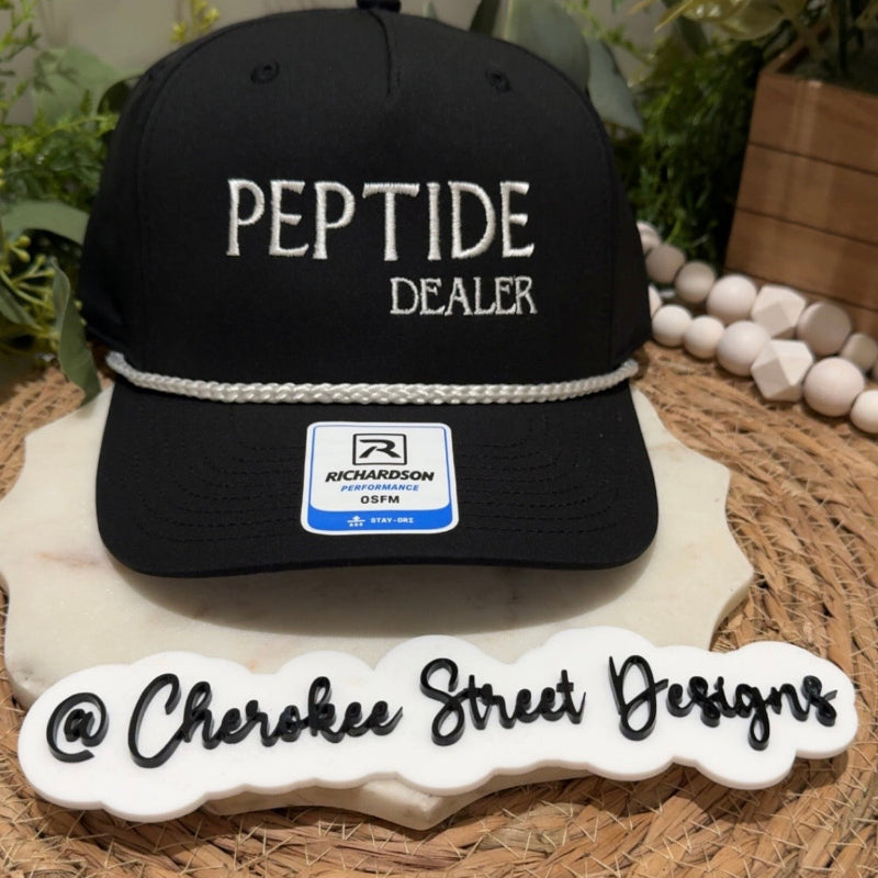 A black baseball cap with 'PEPTIDE DEALER' embroidered in white on the front, displayed on a decorative white stand. The hat has a white rope detail across the brim and a visible Richardson 95FM sticker.
