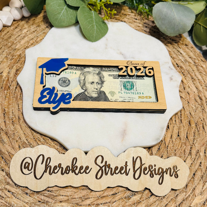 Graduation-themed money card with a blue graduation cap on a dollar bill, placed on a textured surface with greenery.