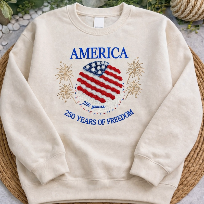 Beige sweatshirt with American flag design and text on a textured surface with plants in the background