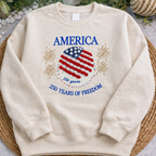 Beige sweatshirt with American flag design and text on a textured surface with plants in the background
