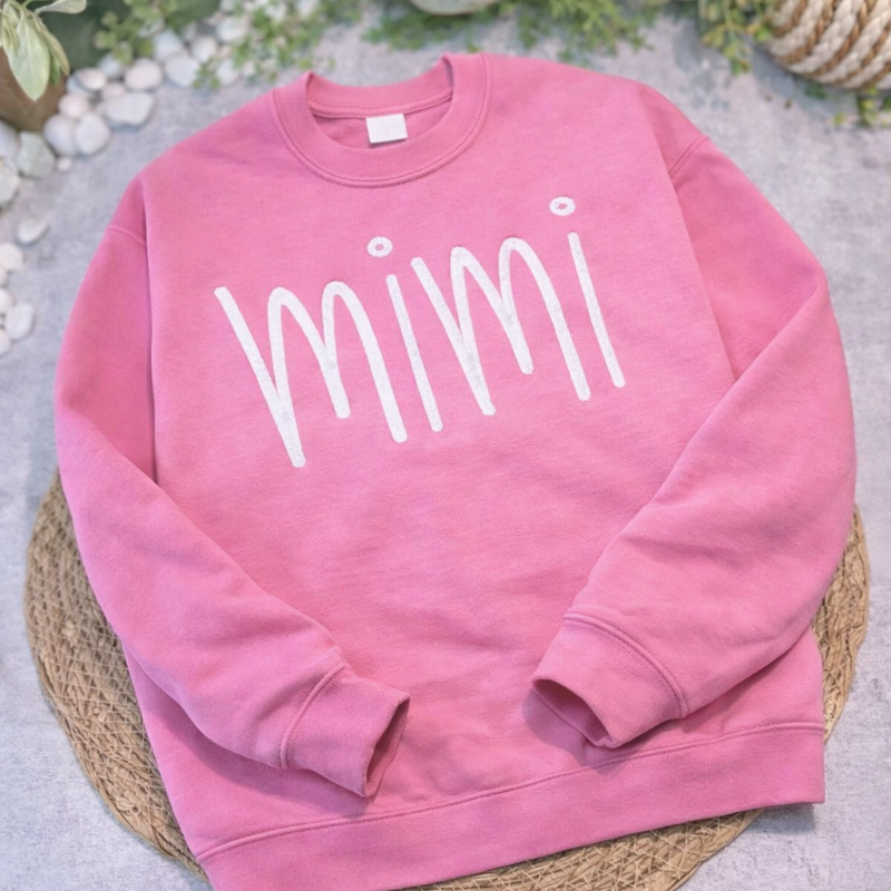Pink sweatshirt with 'mimi' text on a textured surface with greenery