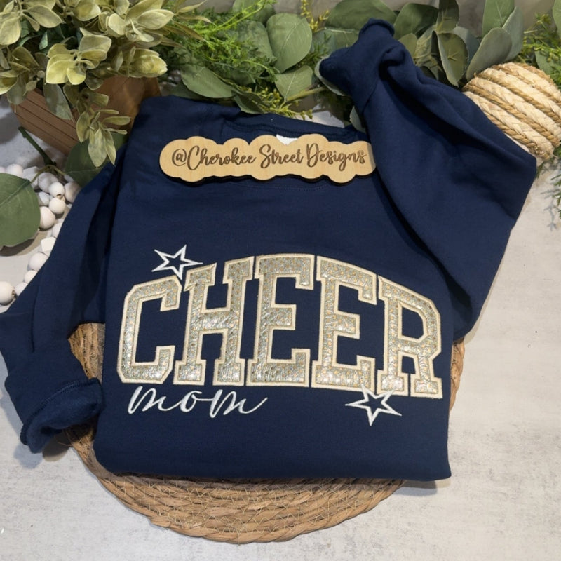 A navy blue sweatshirt laid flat on a woven placemat, with decorative greenery and a marble-patterned surface in the background. The sweatshirt has a tan patch reading 'Charlottes Sweet Designs' and large white 'CHEER mom' lettering with white stars.