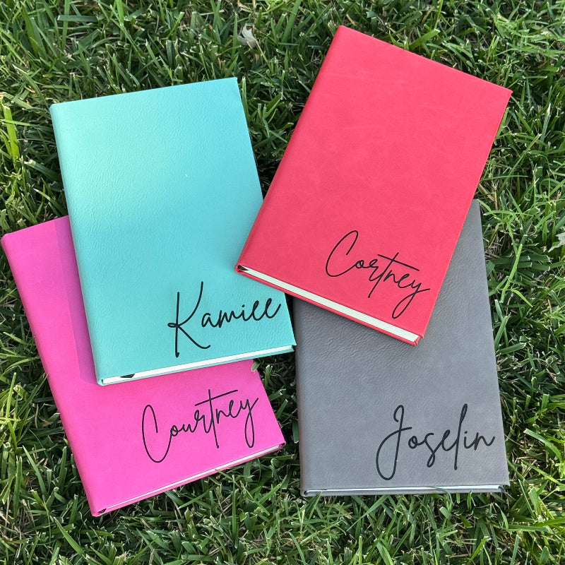 Four hardcover notebooks in red, gray, light blue, and hot pink, arranged on green grass. Each notebook has a black cursive name printed on the cover: Castings, Jocelyn, Kamille, and another Castings.