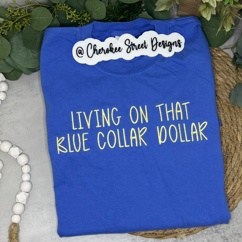 A folded blue graphic t-shirt with yellow text reading 'LIVING ON THAT BLUE COLLAR DOLLAR' 