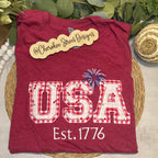Red t-shirt with 'USA Est. 1776' design on a woven mat with greenery in the background