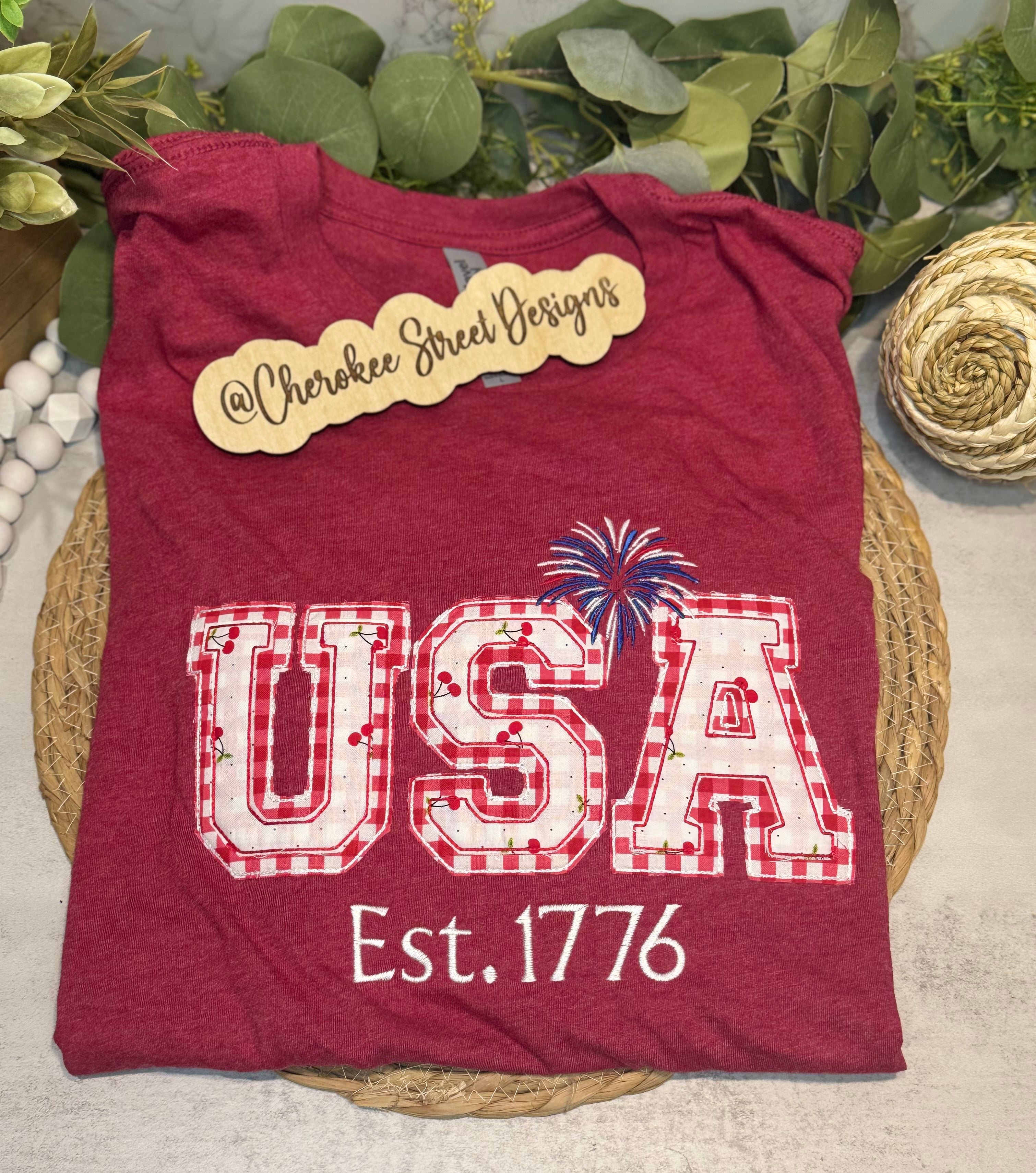 Red t-shirt with 'USA Est. 1776' design on a woven mat with greenery in the background