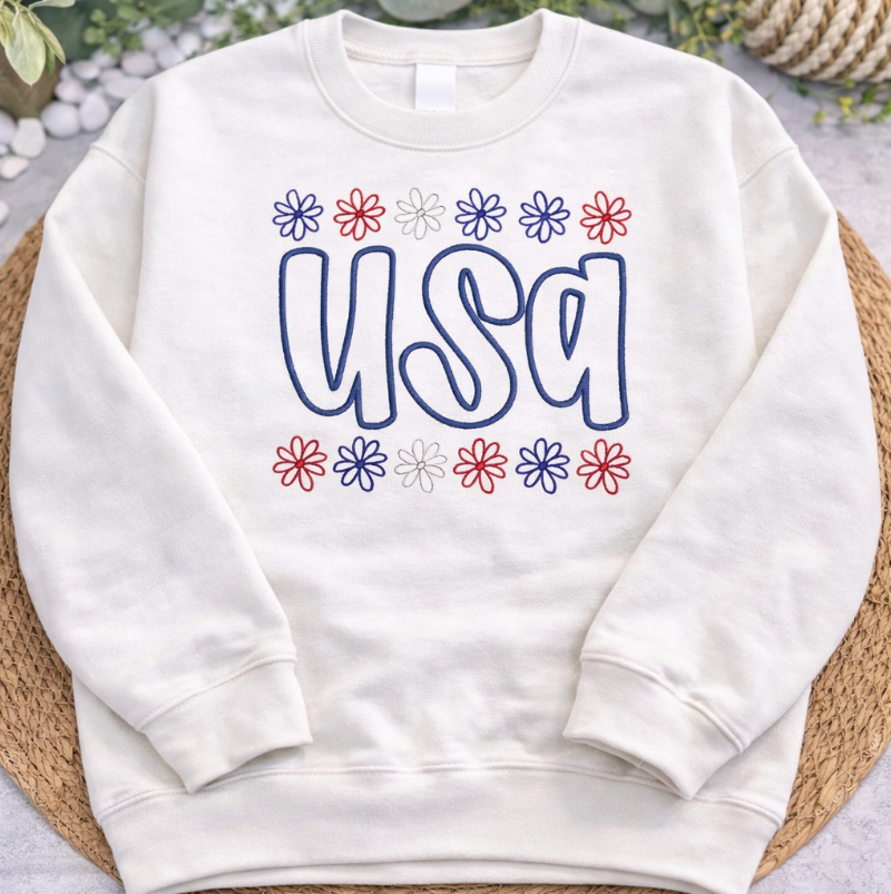 White sweatshirt with 'USA' and floral designs on a textured surface with greenery.