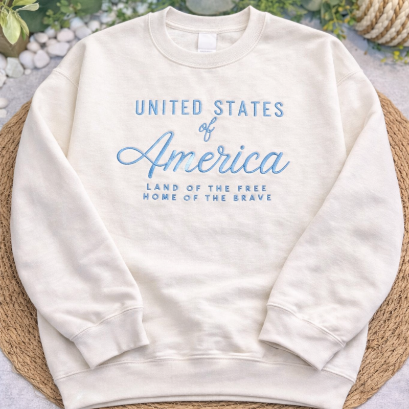 White sweatshirt with 'United States of America' text on a woven mat with greenery in the background
