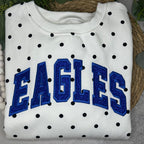A folded white sweatshirt with black polka dots and large blue 'EAGLES' lettering on the front, styled on a woven mat with green leaves in the background.