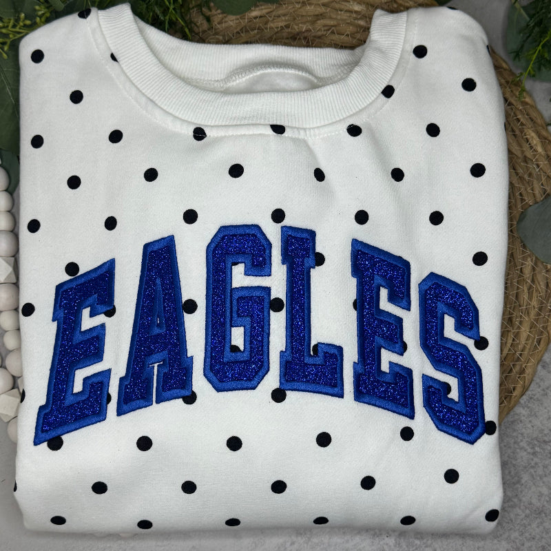 A folded white sweatshirt with black polka dots and large blue 'EAGLES' lettering on the front, styled on a woven mat with green leaves in the background.