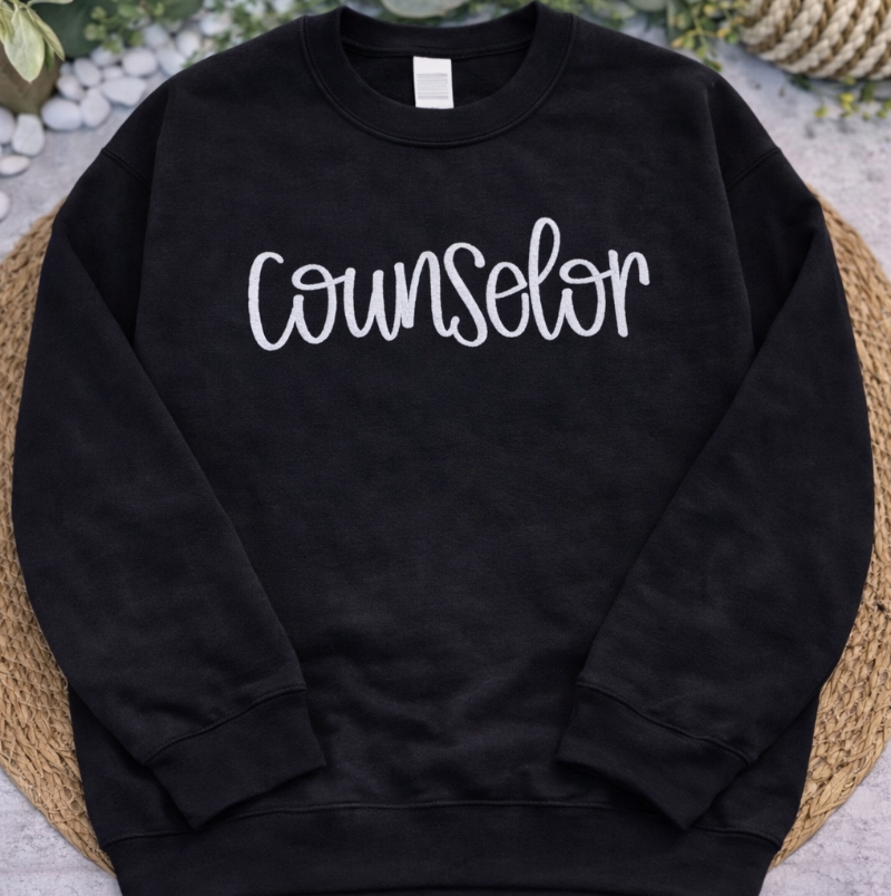 Black sweatshirt with 'Counselor' text on a textured surface with greenery