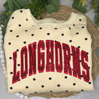 A cream-colored sweatshirt with black polka dots and 'LONGHORNS' in red glittery varsity letters, displayed on a woven placemat with green leaves and white beads.