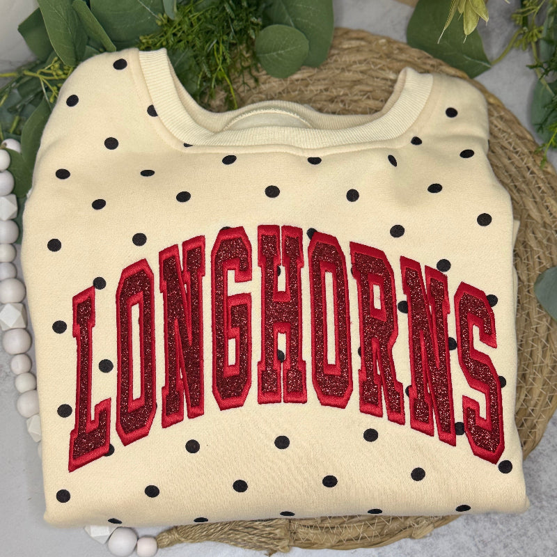 A cream-colored sweatshirt with black polka dots and 'LONGHORNS' in red glittery varsity letters, displayed on a woven placemat with green leaves and white beads.