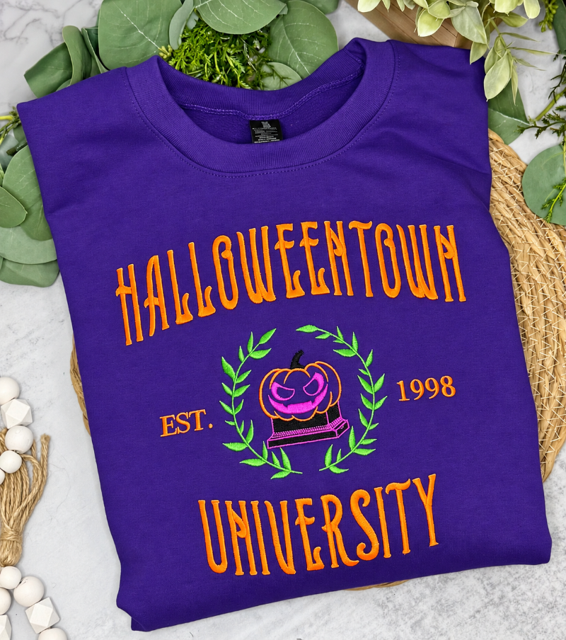 A folded purple t-shirt with a Halloween-themed graphic reading 'HALLOWEENTOWN UNIVERSITY' and 'EST. 1998' around a pumpkin illustration, styled on a light surface with green leaves and woven jute accents.