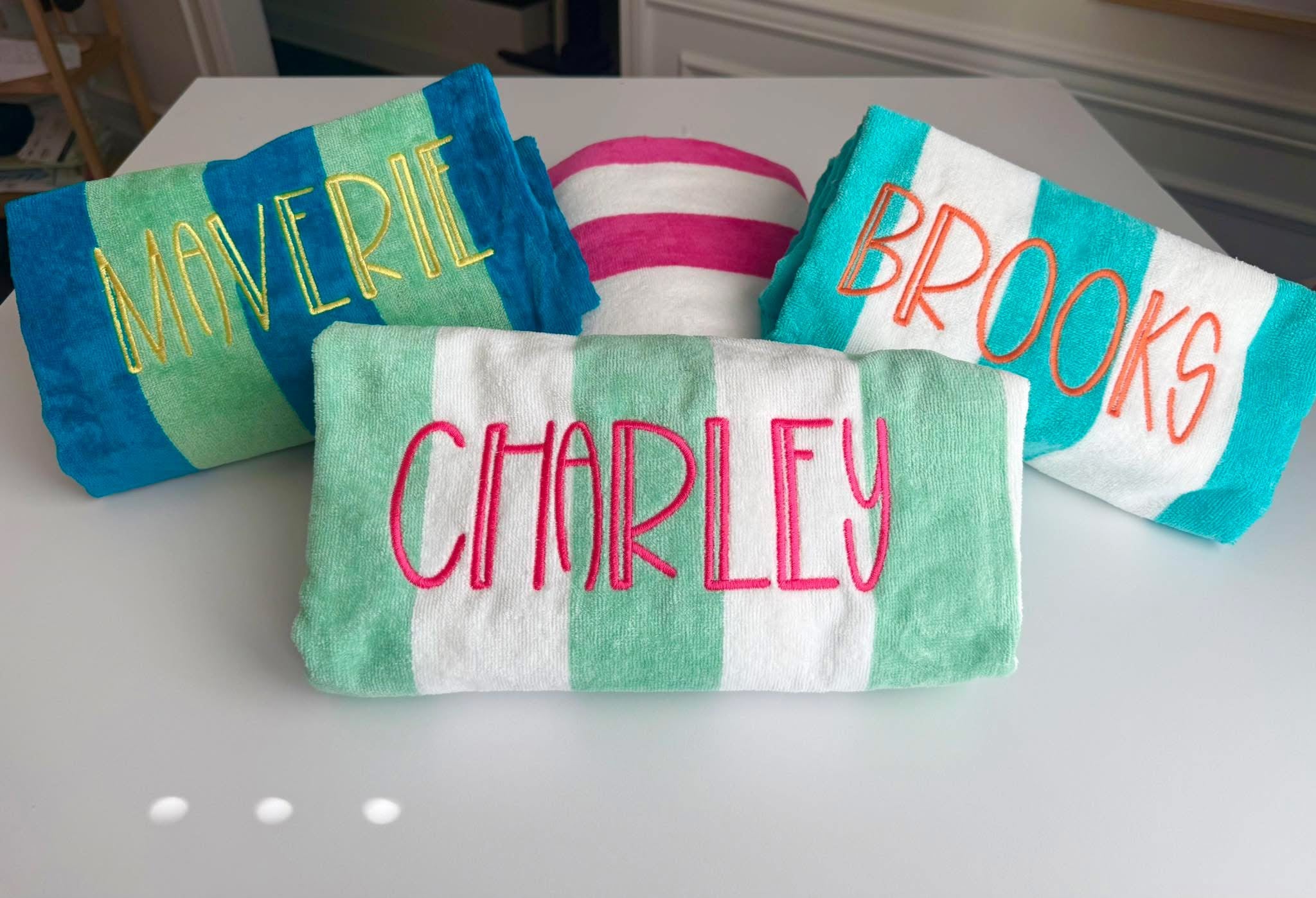 Four beach towels with names embroidered on them, placed on a white surface.