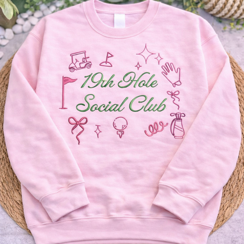 Pink sweatshirt with '19th Hole Social Club' text and graphics on a textured surface with greenery.