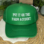 A green trucker-style cap with white embroidered text reading 'PUT IT ON THE FARM ACCOUNT' laid on a woven straw mat.