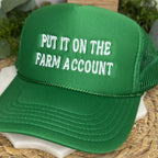 A green trucker hat with white embroidered text reading 'PUT IT ON THE FARM ACCOUNT' resting on a woven straw mat with plants in the background.