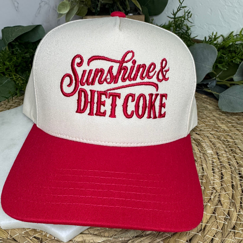 A two-tone trucker cap with an off-white crown and a red brim, featuring the text 'Sunshine & DIET COKE' embroidered in red on the front. The hat is displayed on a woven straw mat with green foliage in the background.