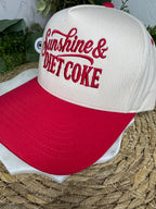 A white and red baseball cap with 'Sunshine & Diet Coke' embroidered on the front, resting on a woven straw surface.