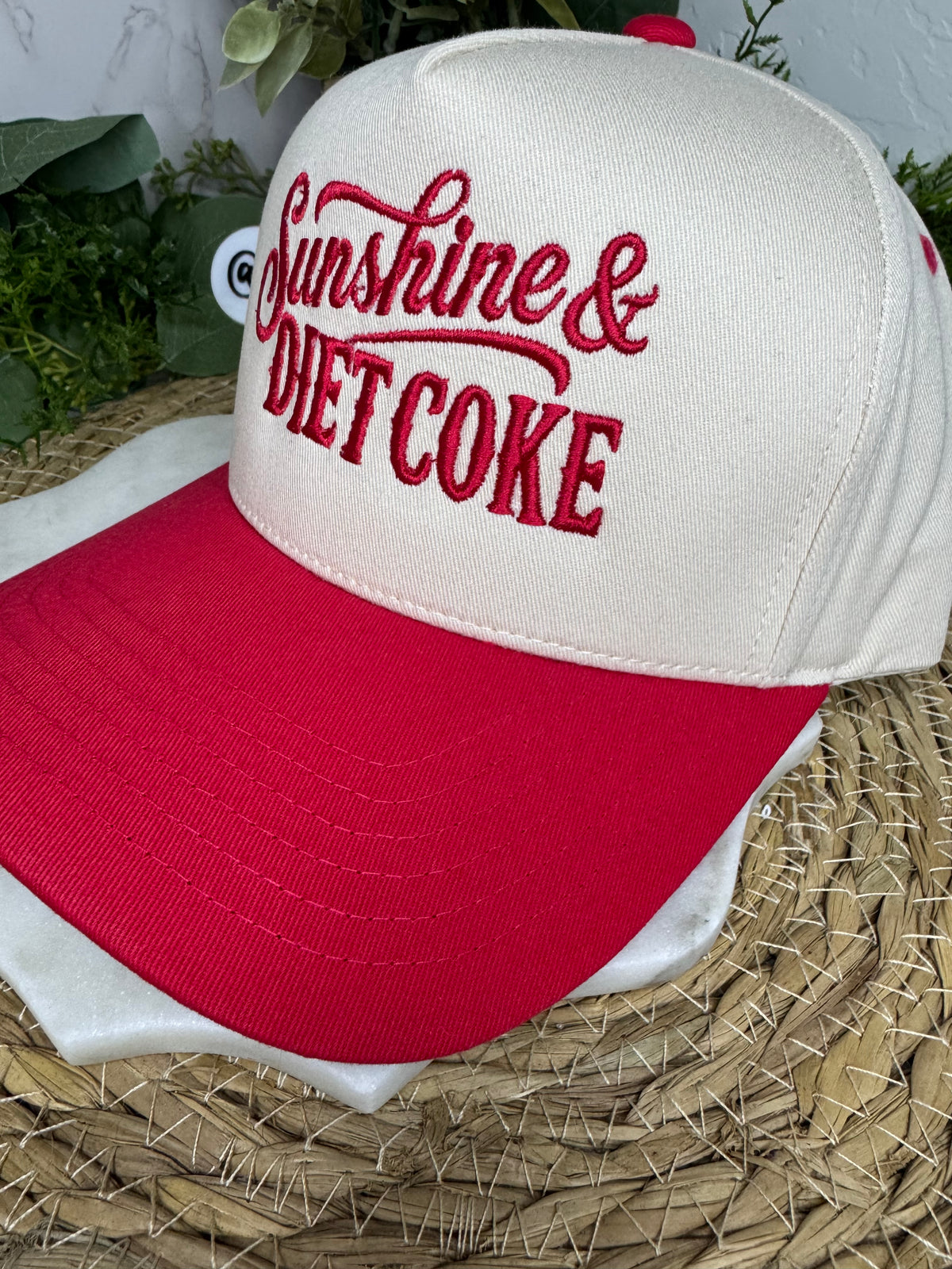 A white and red baseball cap with 'Sunshine & Diet Coke' embroidered on the front, resting on a woven straw surface.