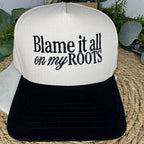 A trucker-style baseball cap with an off-white front panel and black brim, featuring the text 'Blame it all on my ROOTS' embroidered on the front. The cap is displayed on a woven straw mat with green foliage in the background.