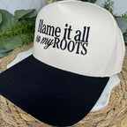 A two-tone trucker-style cap with an off-white crown and a black brim, featuring the text 'Blame it all on my ROOTS' printed in black on the front. The hat is styled on a woven straw mat with green foliage accents.