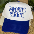 A white and royal blue trucker-style cap with the words 'FAVORITE PARENT' embroidered in blue on the front, resting on a woven mat with blurred greenery in the background.