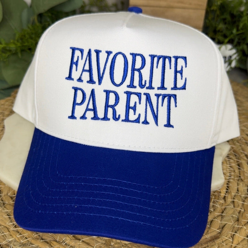 A white and royal blue trucker-style cap with the words 'FAVORITE PARENT' embroidered in blue on the front, resting on a woven mat with blurred greenery in the background.