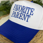 A white and royal blue trucker-style cap with the words 'FAVORITE PARENT' embroidered in blue on the front, resting on a woven straw mat with green plants in the background.