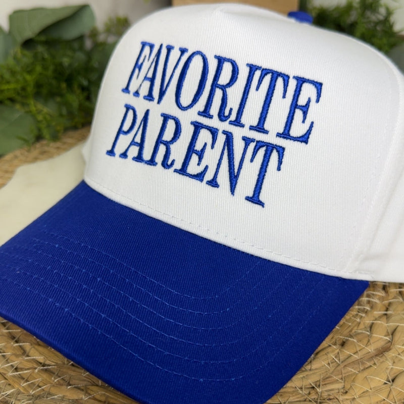 A white and royal blue trucker-style cap with the words 'FAVORITE PARENT' embroidered in blue on the front, resting on a woven straw mat with green plants in the background.