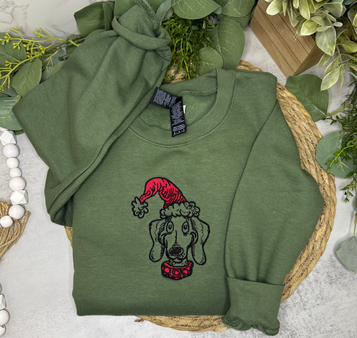 An olive green crewneck sweatshirt laid flat on a woven mat, featuring an embroidered dog wearing a red Santa hat and red collar. The sweatshirt is styled with green leaves, a knotted matching knit piece, and white beads.