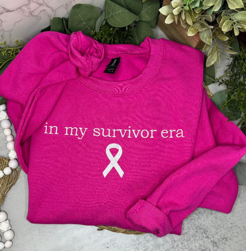 A folded hot pink sweatshirt with the words 'in my survivor era' and a white awareness ribbon printed on the front, styled with green leaves and decorative beads on a light surface.