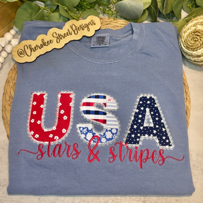 Blue shirt with 'USA stars & stripes' design on a wooden surface with greenery.