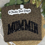 A folded brown leopard-print sweatshirt with 'MOMM'M in large black sequined letters across the front, displayed on a woven mat with greenery and a white bead garland. A white sticker reading '@ Cherokee Street Designs' is placed on the sweatshirt.