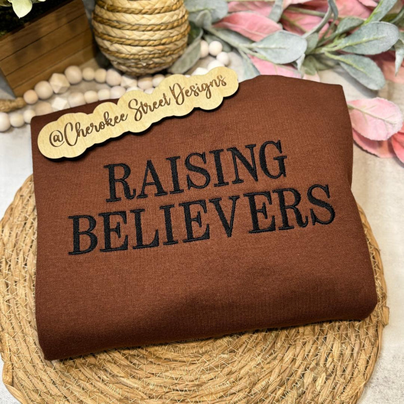 Brown sweatshirt with 'RAISING BELIEVERS' text 