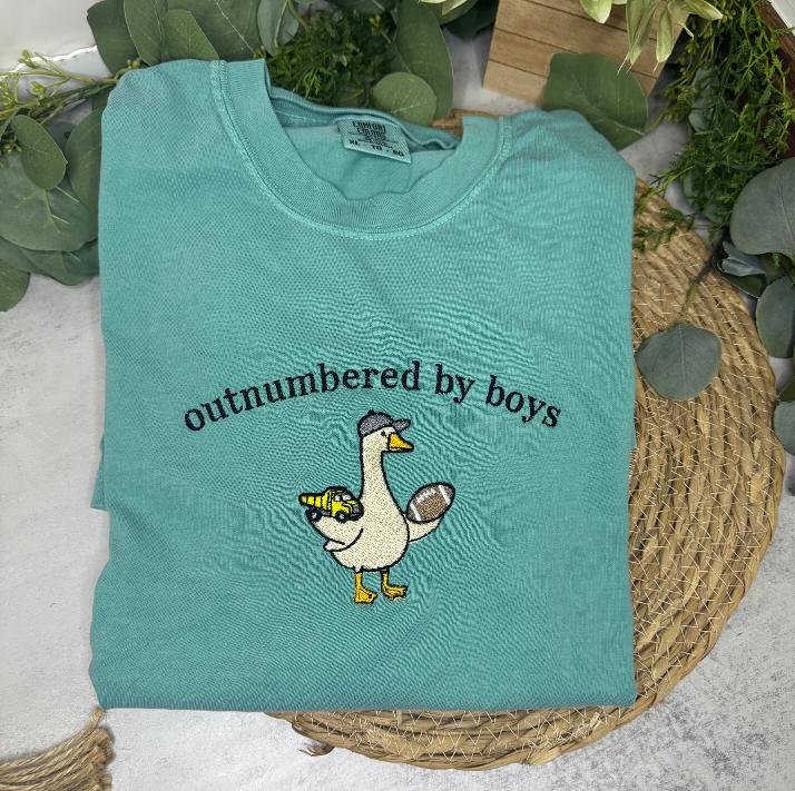 A folded mint green t-shirt with a cartoon goose graphic and the text 'outnumbered by boys' printed on the front, styled on a woven placemat with green leaves.